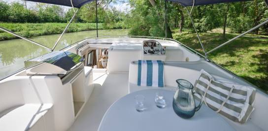 Spacious Mystique sundeck with shaded dining area and helm station on a peaceful river setting.