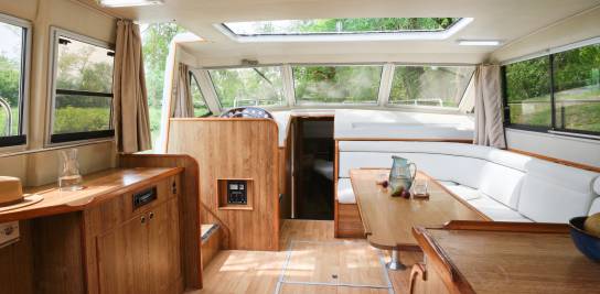 Lichte en luchtige salon van de Grand Classique, met een witte hoekbank, lichtkoepel en een centrale tafel gedekt voor een cruise aan het water.