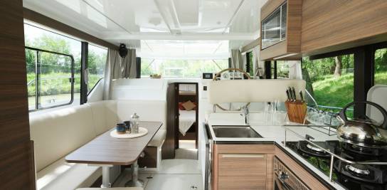 Salon élégant du Liberty avec des fenêtres panoramiques, une grande table en bois, le poste de pilotage et une cuisine complète, le tout dans un espace lumineux et moderne.