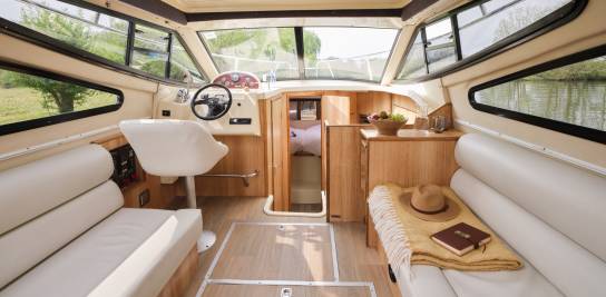 Saloon area aboard the Consul featuring cream seating, helm station, and a wooden cabin entrance, with a hat and book adding a relaxed touch.