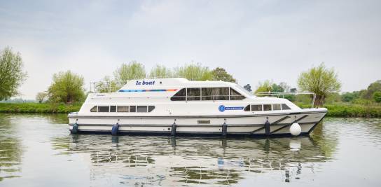 Side view of the Crusader gliding peacefully on a reflective river, with spring-green trees lining the opposite bank and soft clouds overhead.