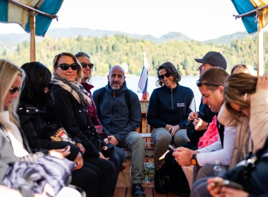 A group of tourists seated on a traditional Pletna boat on Lake Bled, Slovenia, during the AAT Croatia & Slovenia 2025 trip. Passengers are seated facing each other on wooden benches under a striped canopy, with a Slovenian flag visible in the center of the boat. A woman wearing a jacket branded "Europe Express" sits among the group. The scenic backdrop features Lake Bled's forested hillsides and mountains.