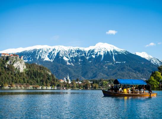 A traditional wooden *pletna* boat with a blue canopy carries passengers across Lake Bled, Slovenia, under a clear blue sky. In the background, Bled Castle sits atop a rocky cliff surrounded by forested hills, with snow-capped Julian Alps mountains stretching across the horizon.