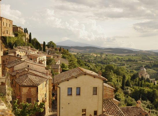 Panoramic view of a historic Tuscan hilltop town, likely Montepulciano, Italy, featuring stone and stucco buildings with terracotta tile roofs cascading down a hillside. A domed church (possibly San Biagio) is visible among lush green trees in the middle distance, with rolling Tuscan countryside, cypress trees, olive groves, and distant mountains stretching to the horizon under a partly cloudy sky.