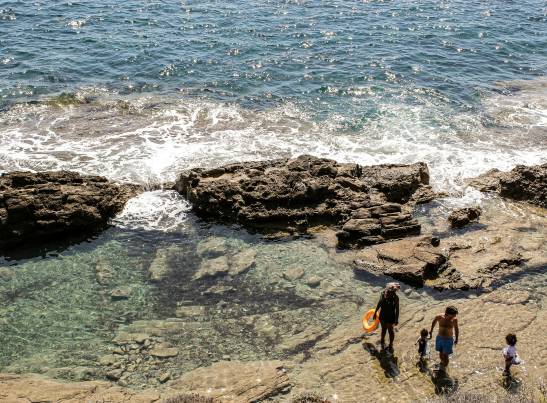Aerial view of a rocky coastal tide pool with clear, shallow water. In the lower right, a family of four — two adults and two young children — explores the rocky shoreline. One adult carries an orange swim ring. Beyond the rocks, ocean waves crash against the shore under bright sunlight.