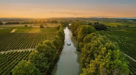 Un bateau vogue vers le coucher de soleil doré sur le Canal du Midi, traversant vignobles et campagne rurale pour un instant photo parfait lors d’un séjour fluvial en France.