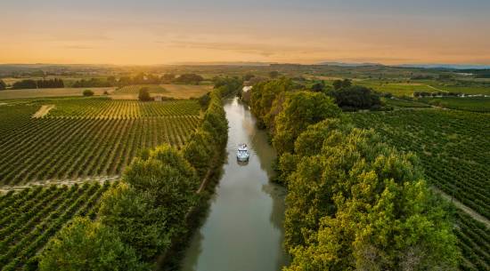 A lone canal boat sails into the golden sunset on the Canal du Midi, cutting through vineyards and rural countryside for a picture-perfect moment on a French boating holiday.