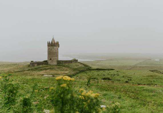 A medieval stone tower castle stands alone on a small hill surrounded by lush green Irish countryside on a misty, overcast day. The circular tower features a pointed turret top and crenellated battlements. Yellow wildflowers are visible in the foreground, while a river or inlet and patchwork fields stretch into the fog in the background. This appears to be Doonagore Castle in County Clare, Ireland.