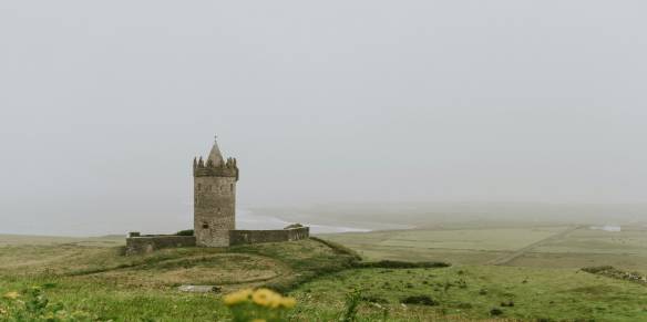 A medieval stone tower castle stands alone on a small hill surrounded by lush green Irish countryside on a misty, overcast day. The circular tower features a pointed turret top and crenellated battlements. Yellow wildflowers are visible in the foreground, while a river or inlet and patchwork fields stretch into the fog in the background. This appears to be Doonagore Castle in County Clare, Ireland.