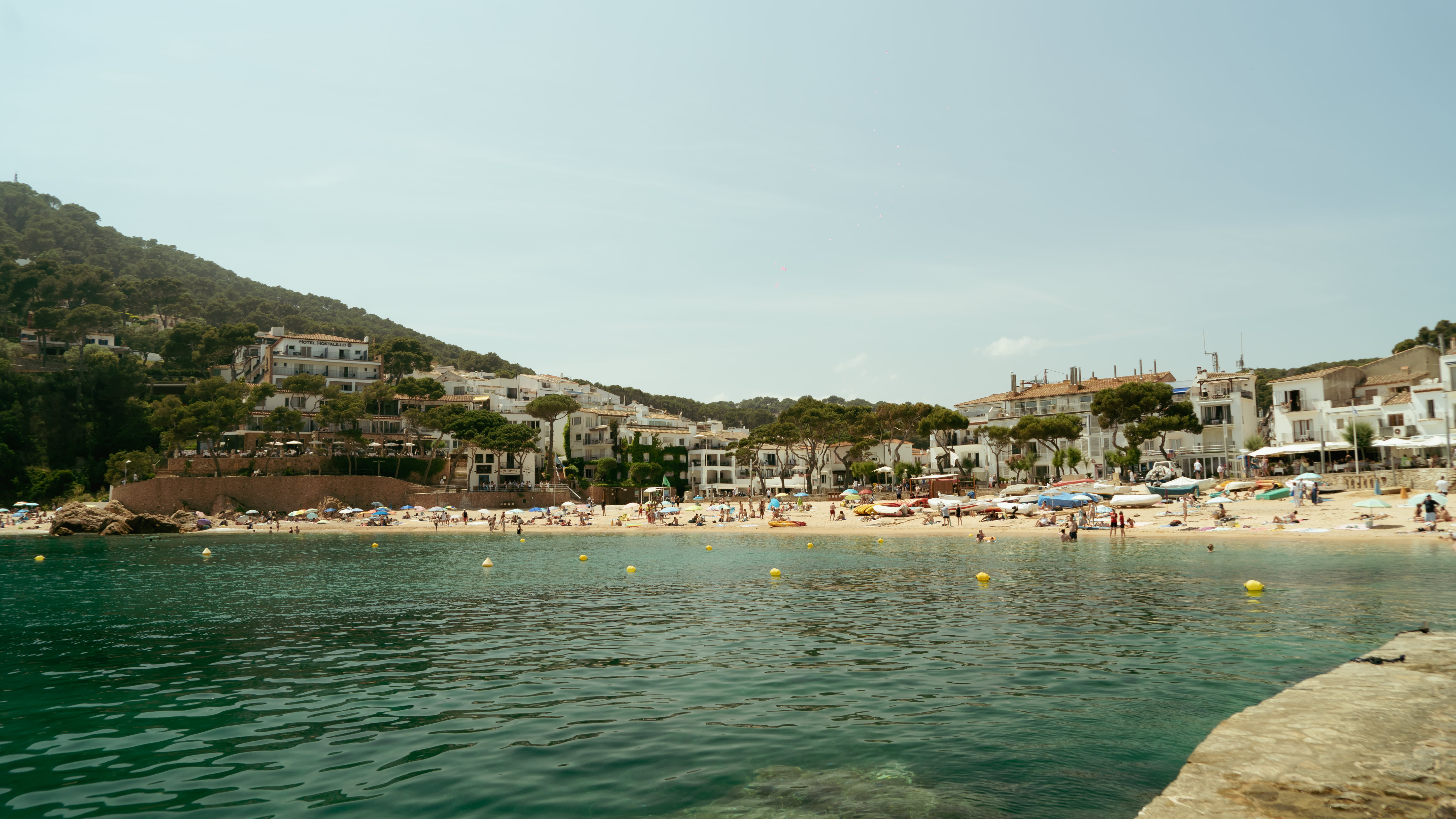 A busy Mediterranean beach cove viewed from the water, with calm turquoise water marked by yellow buoys in the foreground. The sandy beach is lined with sunbathers and colorful umbrellas. Behind the beach, white-washed buildings and hotels are nestled among lush green trees, with a forested hillside rising in the background under a hazy blue sky. The scene resembles a coastal village on the Costa Brava in Spain.