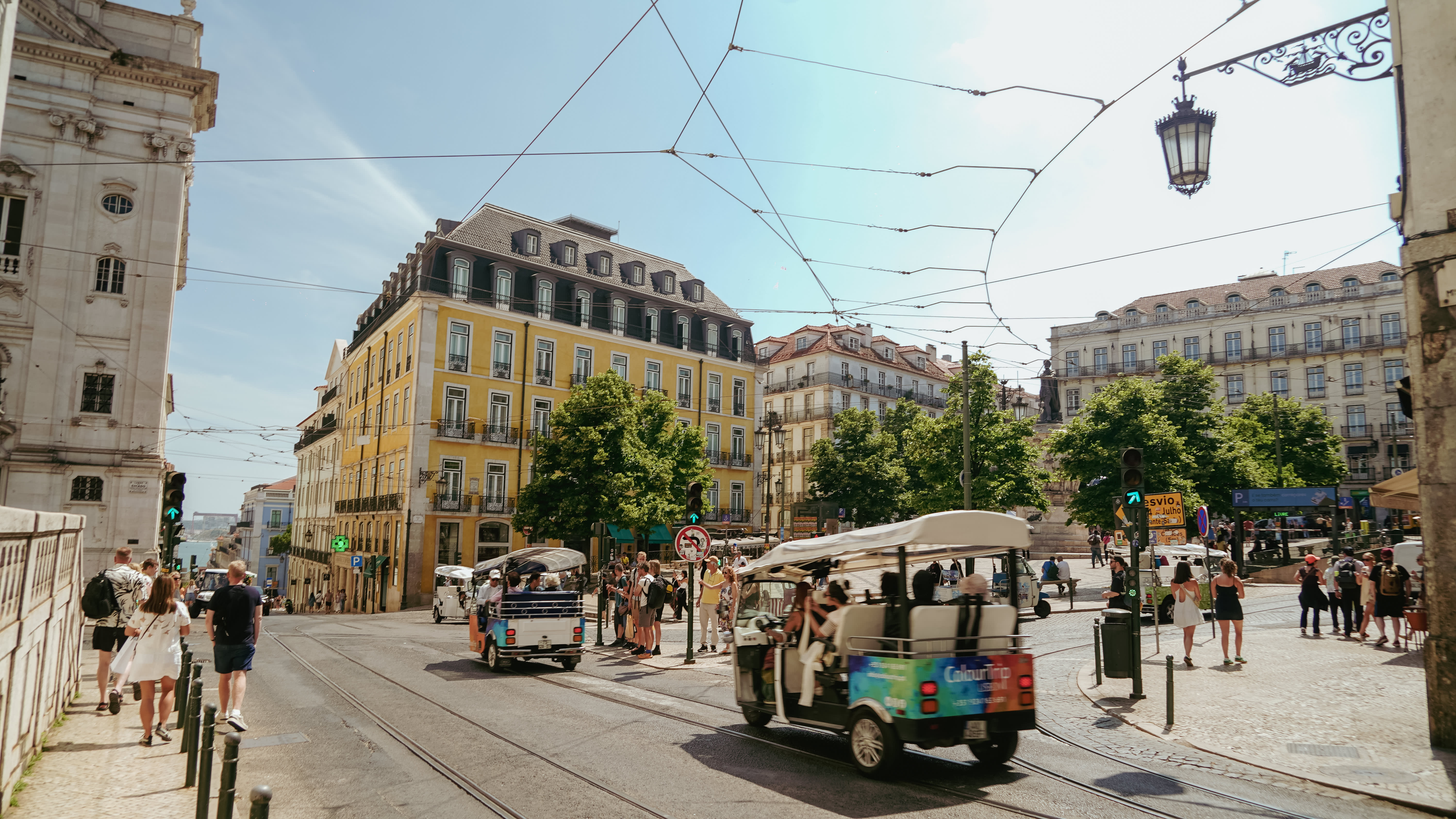 A busy urban square in Lisbon, Portugal, featuring tuk-tuks (small motorized tourist vehicles) navigating cobblestone streets with tram tracks. The scene is framed by classic European architecture, including a prominent yellow building with mansard roof, surrounded by green trees. Overhead tram wires crisscross above the intersection, and a decorative iron street lamp is visible in the upper right. Pedestrians walk along the sidewalks, and a statue is partially visible among the trees in the background. The sky is bright blue with light clouds.