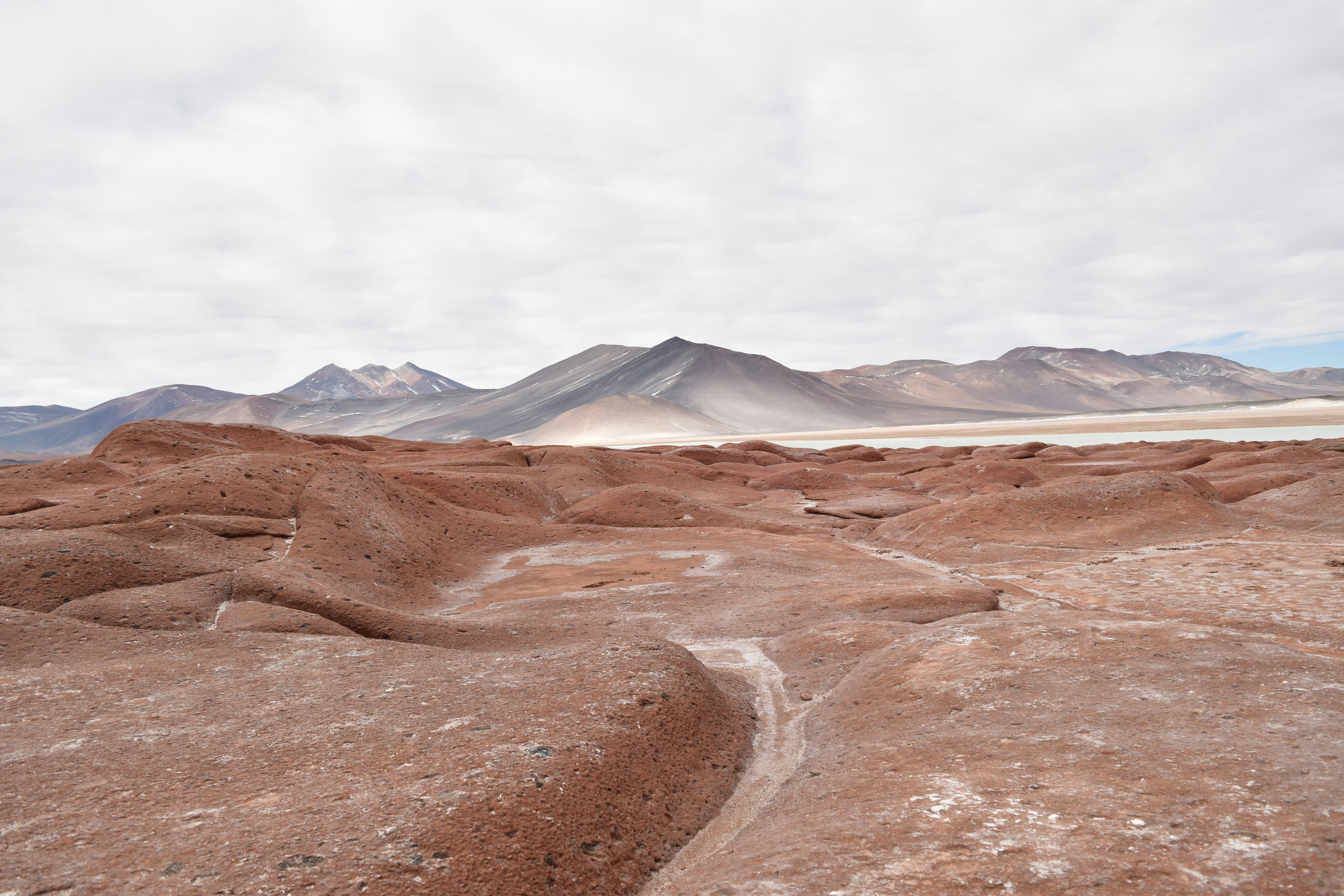 Karge, rotbraune Felslandschaft der Atacama-Wüste in Chile. Im Hintergrund sind dunkle Vulkanberge sowie ein heller Salzsee.