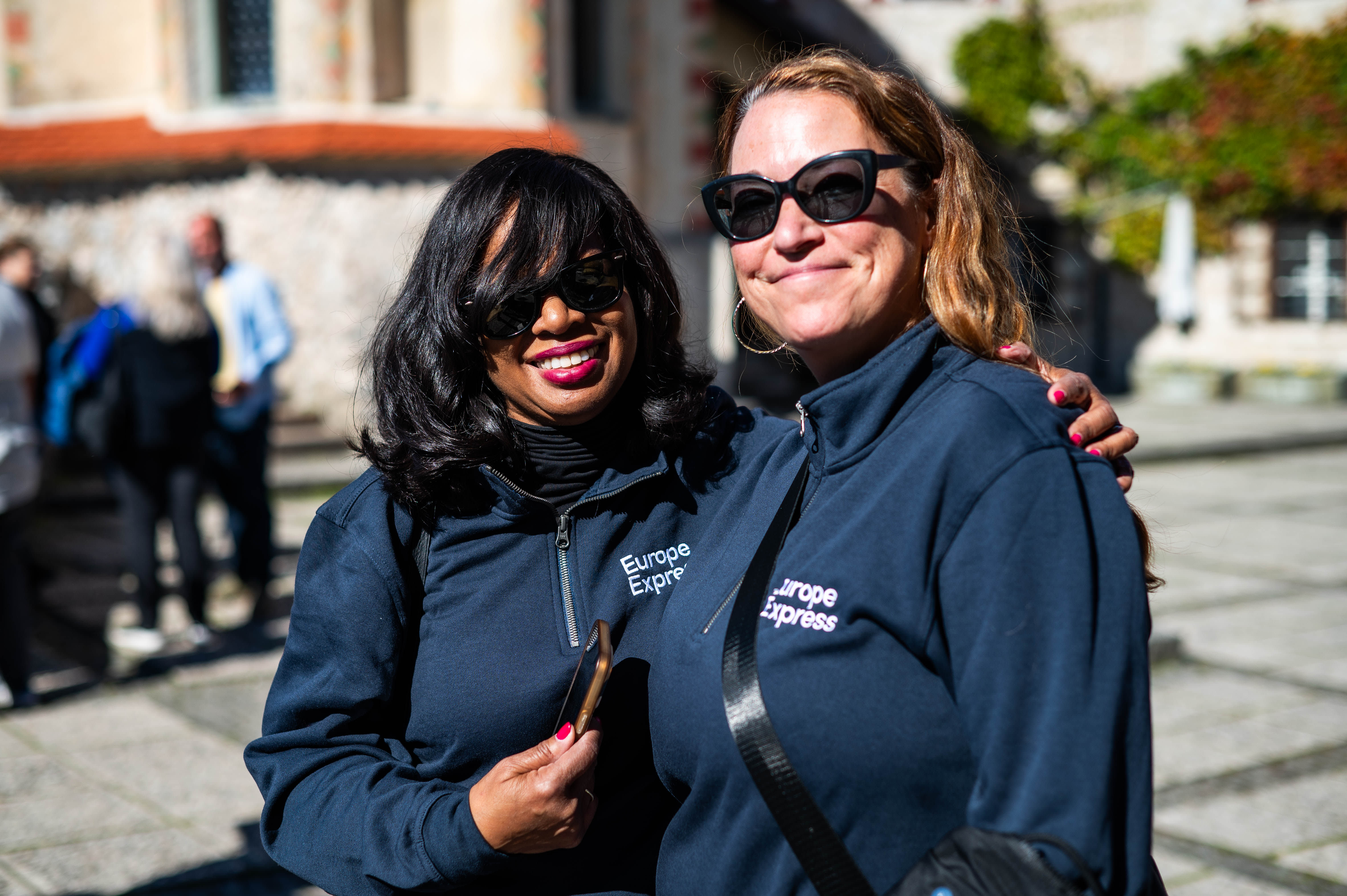 Two smiling women wearing matching navy blue "Europe Express" zip-up jackets and sunglasses pose together outdoors on a sunny day during the AAT Croatia & Slovenia 2025 trip. One woman has her arm around the other's shoulder. A stone plaza and historic building are visible in the blurred background.
