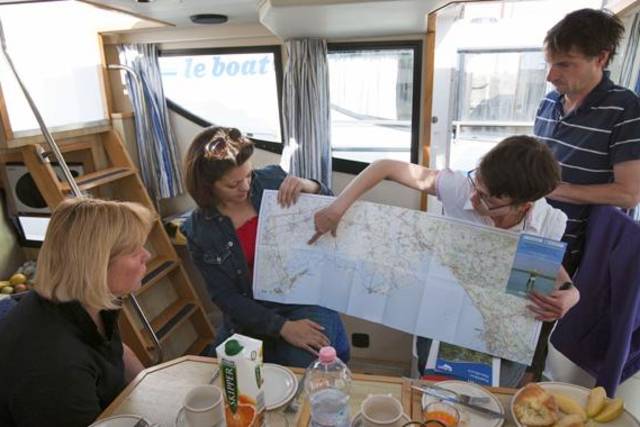 In een Le Boat-cabine zit een groep volwassenen rond de tafel terwijl een teamlid details aanwijst op een grote waterwegenkaart om hen te helpen bij het plannen van hun route.
