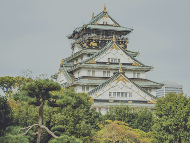 Osaka Castle with green tiled roofs and gold accents rising above lush Japanese garden trees against a gray sky.