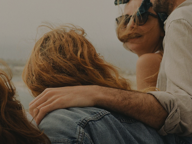 Group of friends outdoors, hair blowing in wind; one wears a red cap with scarf, another has sunglasses, arm around a friend.