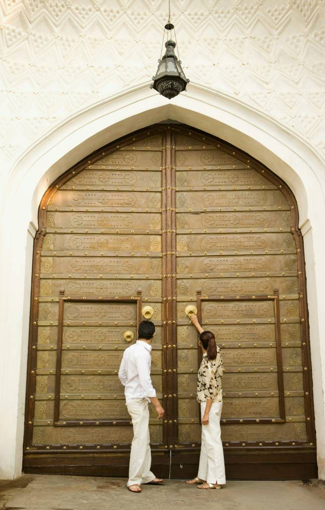 Two people stand before a massive ornate brass door in a white arched entryway, one reaching up to knock.