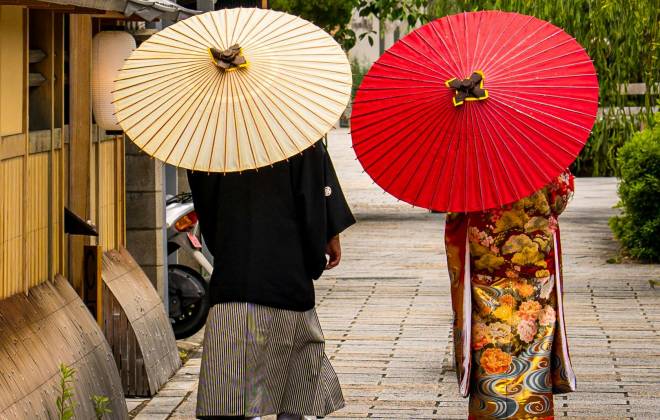 Two people in traditional Japanese kimono walking along a stone path, holding cream and red paper parasols.