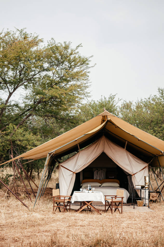 Luxury safari glamping tent in Serengeti savanna with a dining table, chairs, and bed visible, surrounded by African bush trees.