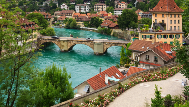 Aerial view of a historic European city, likely Bern, Switzerland, featuring a stone arch bridge spanning the turquoise Aare River. The scene includes traditional buildings with red-tiled roofs, lush green trees, a flowering garden path in the foreground, and a dense residential neighborhood climbing the hillside in the background.