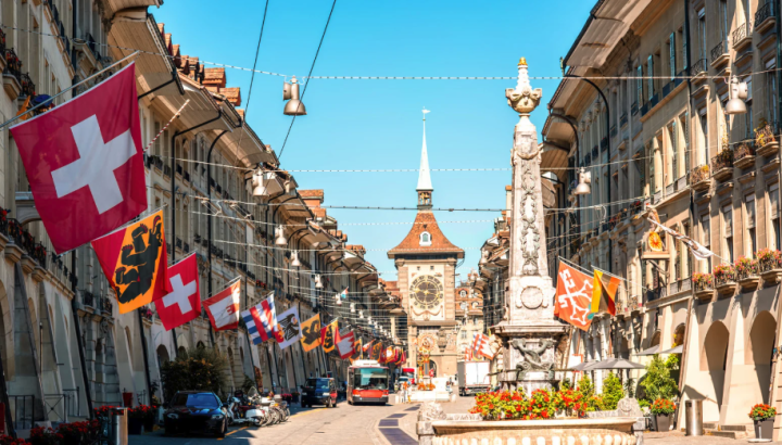 A sunny street scene in Bern, Switzerland's old town (Kramgasse), featuring the iconic Zytglogge (Clock Tower) at the end of the pedestrian street. The street is decorated with numerous Swiss national flags (red with white cross) and cantonal flags hanging between historic sandstone buildings. In the foreground stands an ornate medieval fountain (Simsonbrunnen). A red tram/bus is visible near the clock tower, and flower boxes with red blooms decorate the buildings under a clear blue sky.