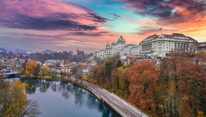 Aerial view of Bern, Switzerland, featuring the Swiss Federal Palace (Bundeshaus) with its distinctive green dome and Swiss flags, situated on a hill above the Aare River. The scene is captured during sunset with dramatic pink and purple clouds. Autumn foliage in orange and red tones surrounds the historic buildings, and a riverside road runs along the water's edge.