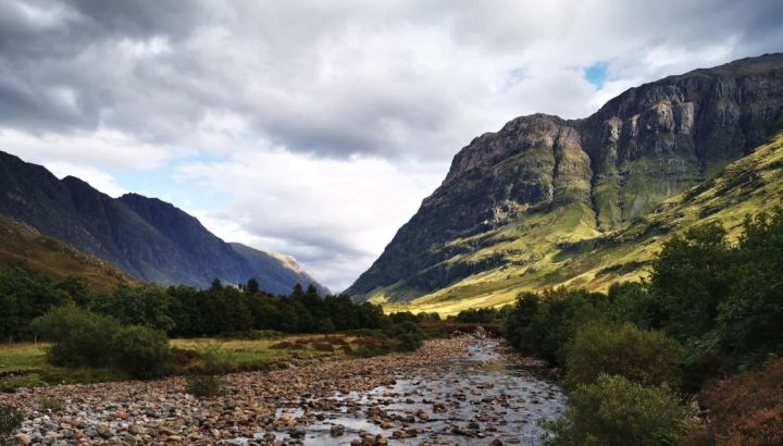 A shallow, rocky river flows through a dramatic Scottish Highland valley, likely Glencoe, with steep, rugged mountains rising on both sides. The right mountain is sunlit, revealing vivid green slopes, while the left remains in shadow. Trees and shrubs line the riverbanks, and a cloudy sky with patches of blue stretches overhead.