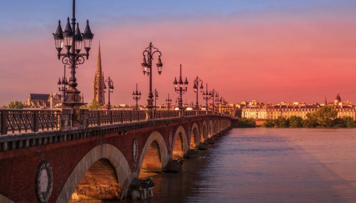**Pont de Pierre (Stone Bridge) in Bordeaux, France at sunset** A historic multi-arched stone bridge spanning the Garonne River, lined with ornate black iron lampposts. The warm pink and purple sunset sky reflects on the calm river water. In the background, the Bordeaux city skyline is visible, including the spire of the Basilica of Saint-Michel and classic Haussmann-style buildings along the riverbank.