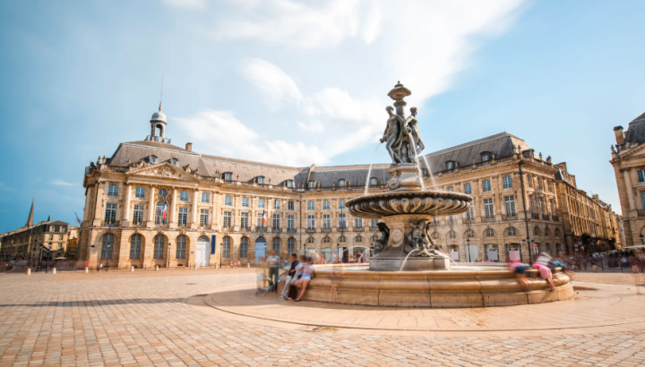 The Fountain of the Three Graces at Place de la Bourse in Bordeaux, France. The ornate baroque fountain features bronze sculptural figures in the center of a large open cobblestone plaza. Behind it stands the grand 18th-century Palais de la Bourse, a curved neoclassical limestone building with mansard roofs and ornate architectural details. Several blurred figures of people are visible near the fountain base, suggesting a long-exposure photograph taken on a sunny day with a partly cloudy blue sky.