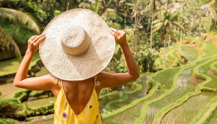 Is Indonesia safe? Beautiful young lady in shine through dress touch straw hat. Girl walk at typical Asian hillside with rice farming, mountain shape green cascade rice field terraces paddies. Ubud, Bali, Indonesia