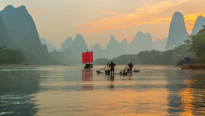 Fisherman stands on traditional bamboo boats at sunrise (boat with a red sail in the background)