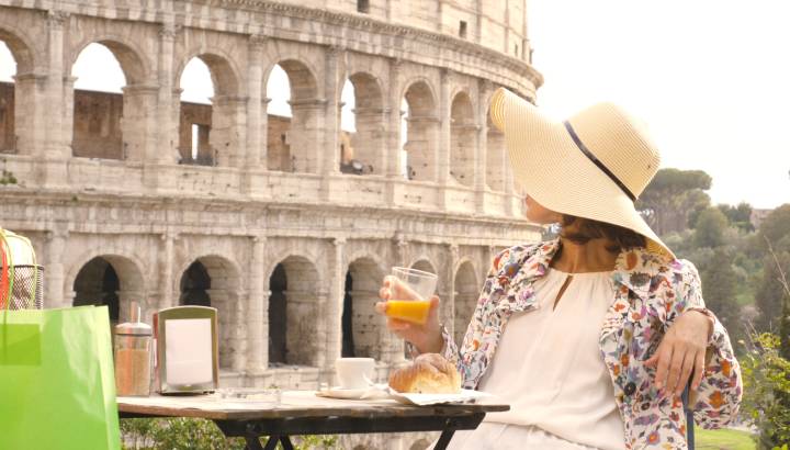 Happy young woman tourist drinking coffee and juice with cornetto at the table outside a bar restaurant in front of the Colosseum in Rome