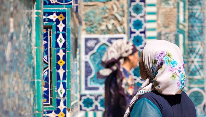 Verschwommen im Vordergrund zwei traditionell gekleidete Frauen vor dem Hintergrund des historischen Friedhofs Shahi Zinda in Samarkand, Uzbekistan
