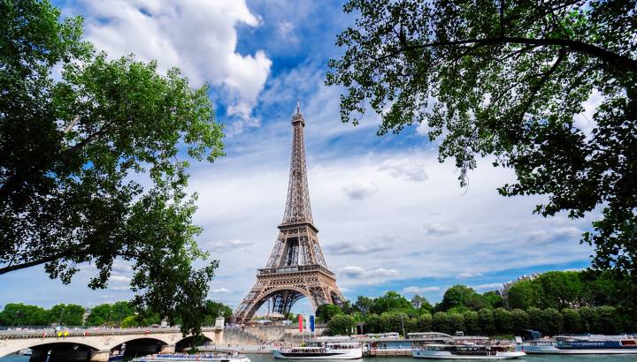 Eiffel Tower rising above the Seine River, framed by green trees, with tour boats and a bridge in Paris, France.