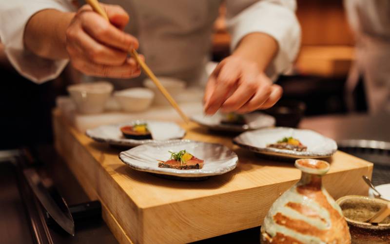 Chefs using chopsticks to plate elegant Japanese dishes with fish and garnish on ceramic plates atop a wooden board.