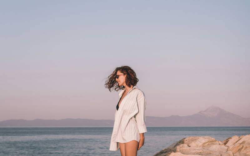 Woman in white linen shirt and shorts standing barefoot on coastal rocks, hair blowing in wind, calm sea and mountains behind her.