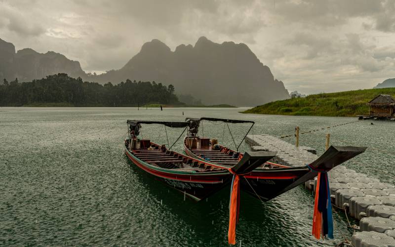 Two traditional Thai longtail boats docked at a floating pier on a misty lake, with dramatic limestone mountains in the background.
