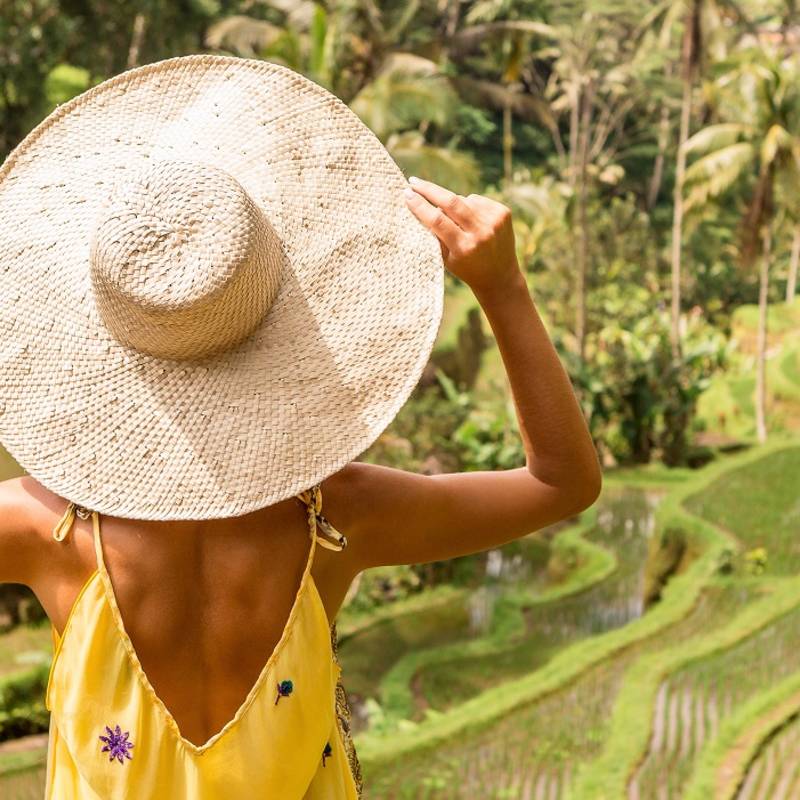 Is Indonesia safe? Beautiful young lady in shine through dress touch straw hat. Girl walk at typical Asian hillside with rice farming, mountain shape green cascade rice field terraces paddies. Ubud, Bali, Indonesia