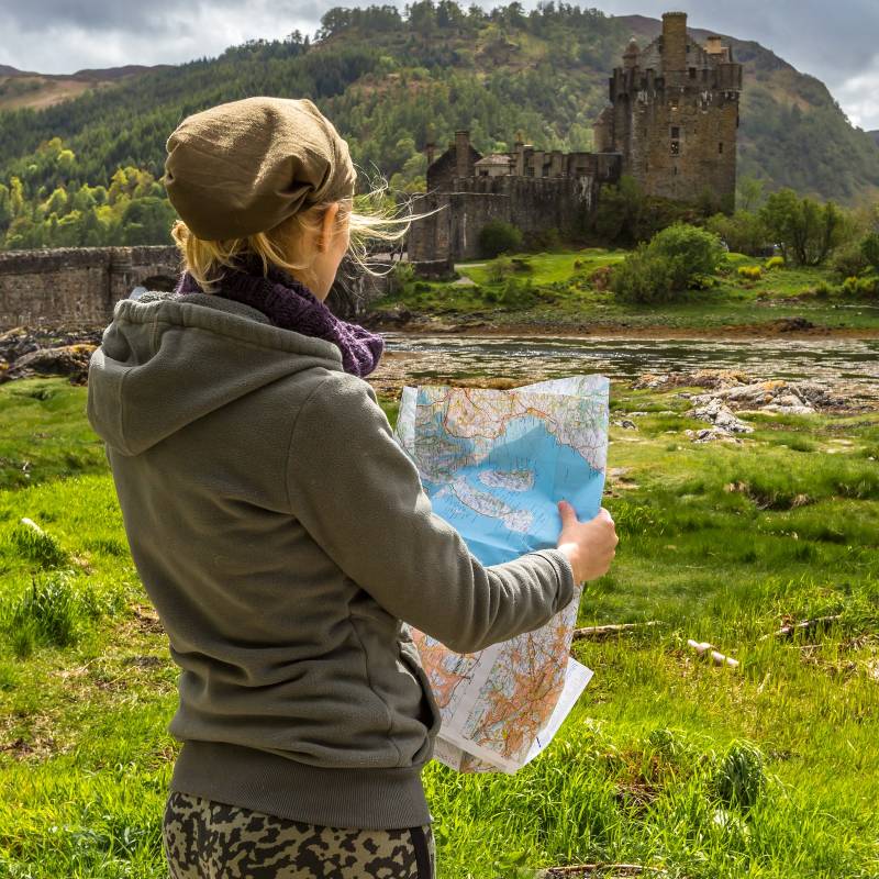 Young woman tourist looks at a road map. In background the famous and spectacular Eilean Donan Castle in Dornie, Kyle of Lochalsh, Scotland, United Kingdom