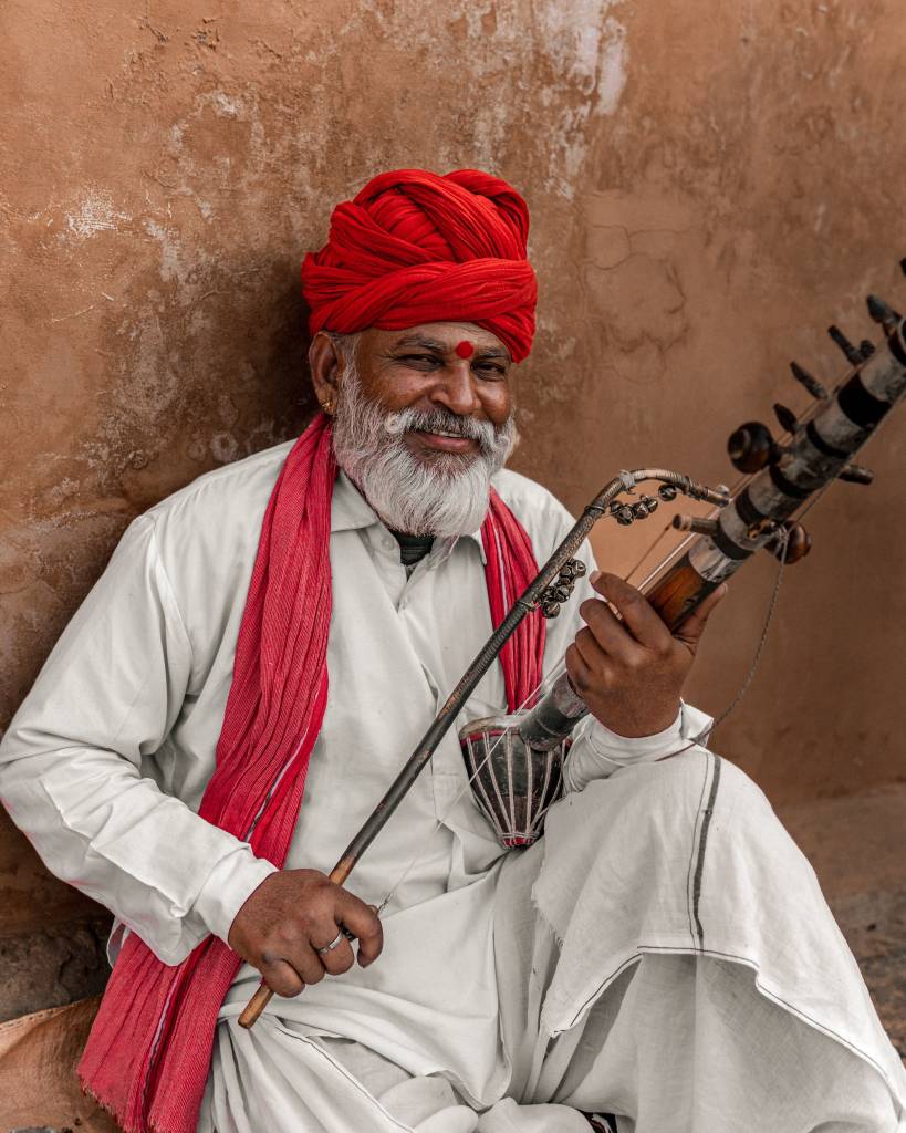 Smiling elderly Indian man with white beard, red turban and scarf, playing a traditional sarangi instrument against a clay wall.