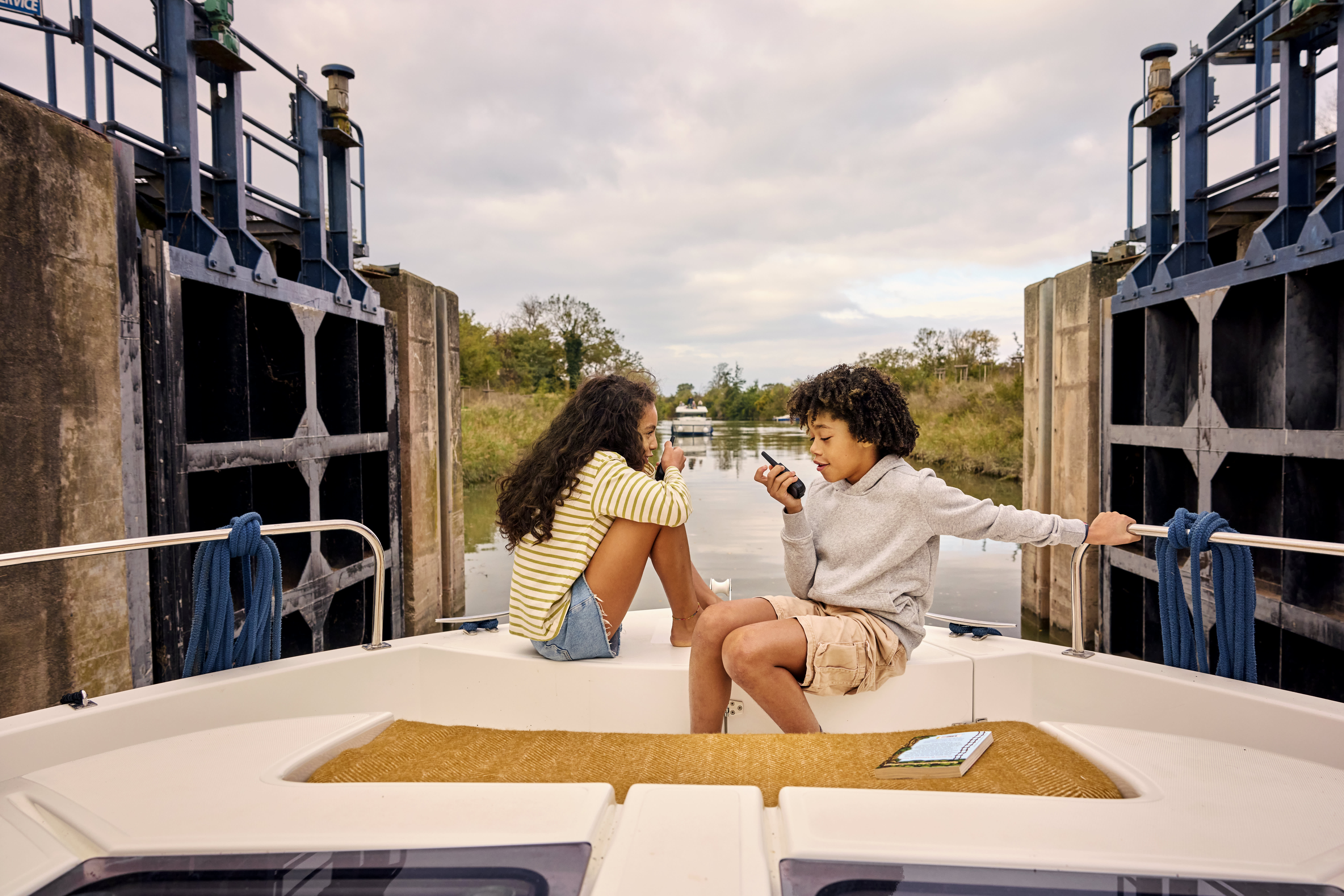 Two children relaxing on the front deck of a boat while passing through a canal lock during a family boating holiday.