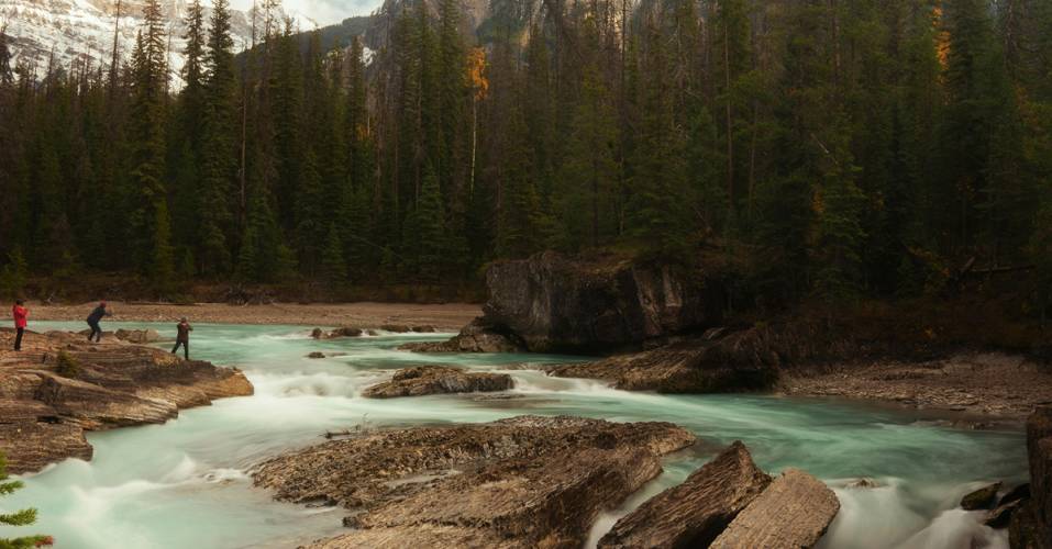 A turquoise glacial river flowing over rocky terrain in a Canadian wilderness setting, likely in the Rocky Mountains. Three visitors stand on the rocky riverbank observing the rushing water, surrounded by dense evergreen forest with snow-capped mountain peaks visible in the background. The autumn foliage adds touches of yellow among the conifers.