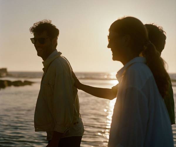 Couple near the ocean at sunset; woman places hand on smiling man's back, both wearing sunglasses, warm golden light.