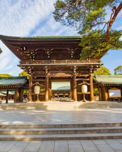 Meiji Shrine's grand wooden torii gate with paper lanterns, surrounded by lush trees under a blue sky in Tokyo, Japan.