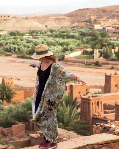 The girl-traveler in a hat is standing on a high wall with arms outstretched in the background of the city in the desert of Ait BenHaddou, Morocco, Africa