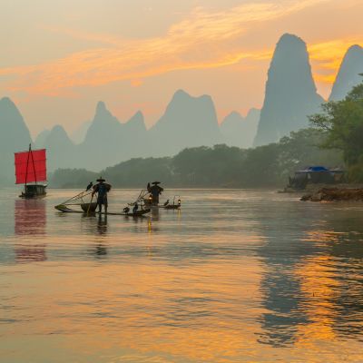 Fisherman stands on traditional bamboo boats at sunrise (boat with a red sail in the background)