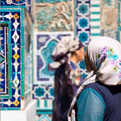Verschwommen im Vordergrund zwei traditionell gekleidete Frauen vor dem Hintergrund des historischen Friedhofs Shahi Zinda in Samarkand, Uzbekistan