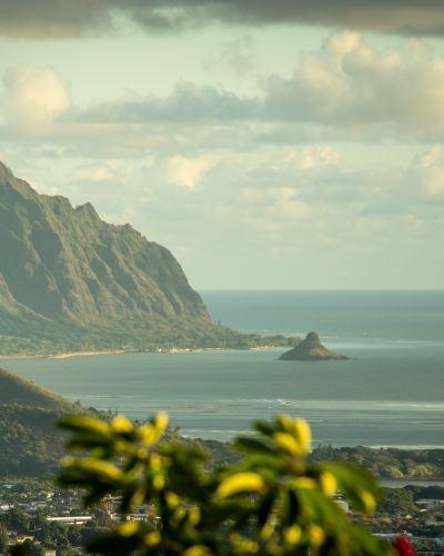 Dramatic Ko'olau Mountain Range cliffs rise above Oahu's coastline, with lush greenery, a bay, and small island visible below.