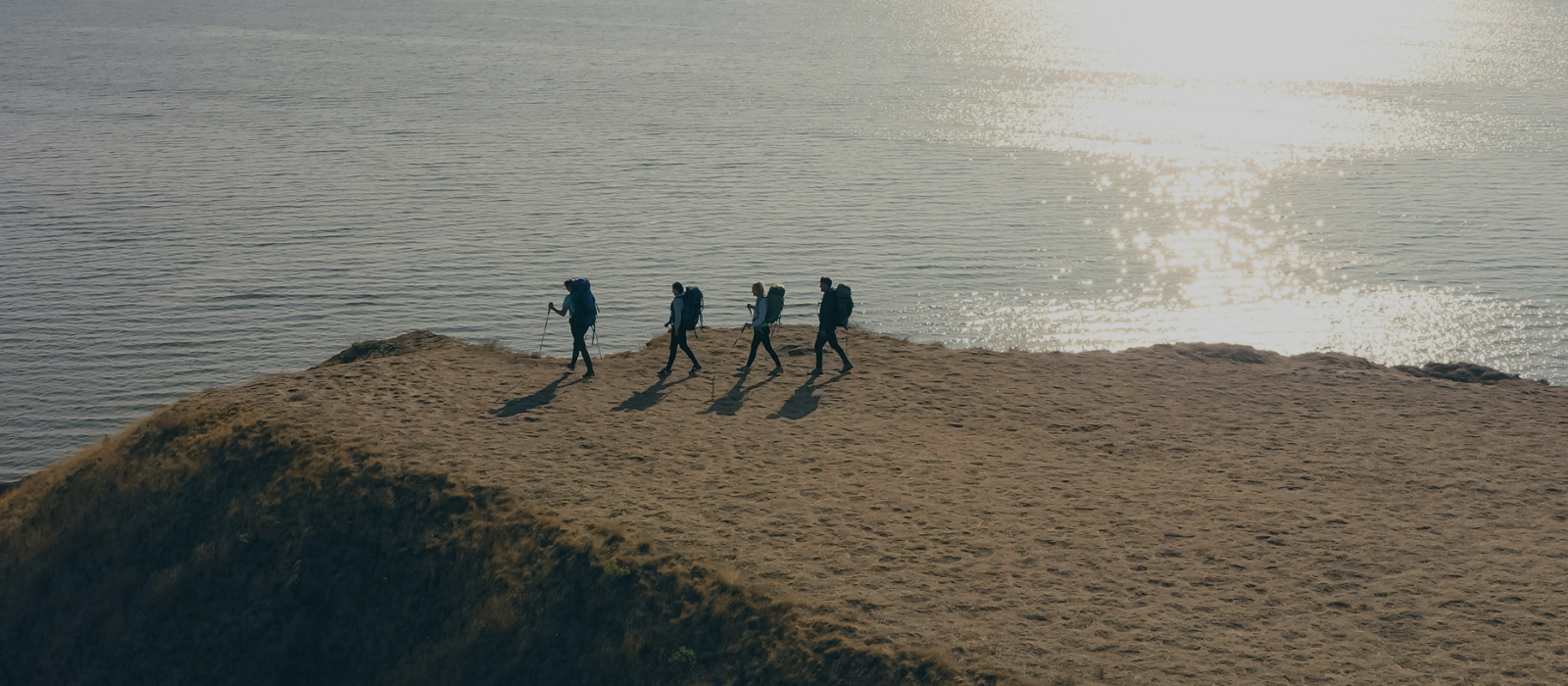 Four hikers with backpacks and trekking poles walking along a coastal ridge with sunlit water in the background.