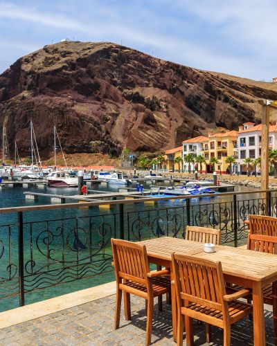 Chairs and tables of local restaurant in harbour with boats and yachts and colorful houses, Madeira island, Portugal